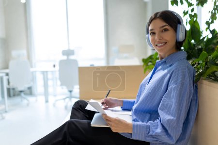 Foto de Señora sonriente en los auriculares con un bolígrafo y documentos en sus manos sentadas en el espacio de coworking - Imagen libre de derechos