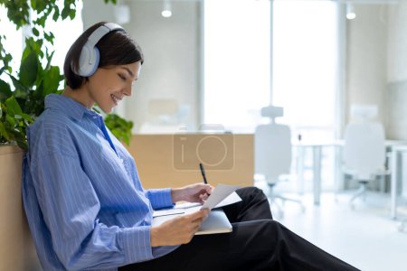 Foto de Vista lateral de una joven alegre en los auriculares con una pluma y papeles en las manos sentadas en el interior - Imagen libre de derechos