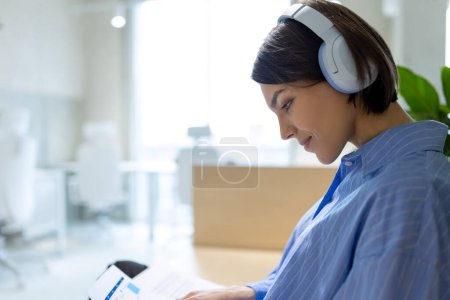 Photo for Side view of a focused young Caucasian woman in the wireless headphones looking through documents - Royalty Free Image
