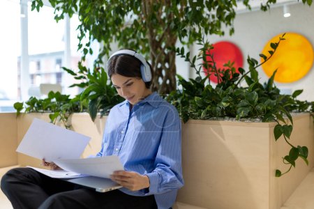 Foto de Mujer enfocada en los auriculares inalámbricos sosteniendo papeles en sus manos en un espacio de coworking - Imagen libre de derechos