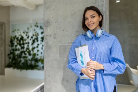 Photo for Smiling happy young woman with business documents and the laptop in her hands leaning against the wall - Royalty Free Image