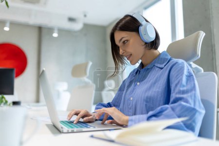 Téléchargez les photos : Employée concentrée dans les écouteurs sans fil assis au bureau travaillant sur l'ordinateur portable - en image libre de droit