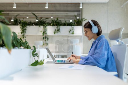 Téléchargez les photos : Vue latérale d'un employé joyeux de l'entreprise dans les écouteurs regardant l'écran de l'ordinateur portable tout en écrivant quelque chose - en image libre de droit
