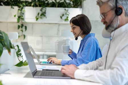 Foto de Vista lateral de los trabajadores de oficina en los auriculares sentados en el escritorio trabajando en los ordenadores portátiles - Imagen libre de derechos
