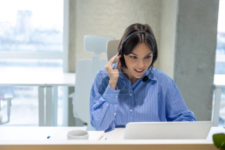 Foto de Trabajador de oficina sonriente en los auriculares sentados en el escritorio mirando la pantalla del portátil - Imagen libre de derechos