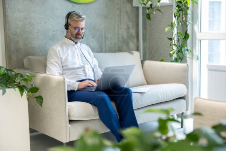 Téléchargez les photos : Homme d'affaires mature concentré dans le casque assis sur le canapé tapant sur l'ordinateur portable - en image libre de droit