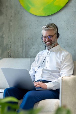 Téléchargez les photos : Joyeux entrepreneur masculin dans le casque assis sur le canapé tapant sur l'ordinateur portable - en image libre de droit