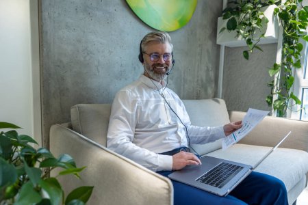 Téléchargez les photos : Homme d'affaires joyeux dans le casque assis sur le canapé avec des documents à la main tapant sur son ordinateur portable - en image libre de droit