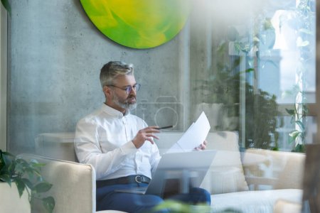 Téléchargez les photos : Entrepreneur masculin concentré sérieux assis sur le canapé avec l'ordinateur portable regardant à travers les papiers dans sa main - en image libre de droit