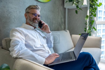 Téléchargez les photos : Homme entrepreneur mature souriant avec l'ordinateur portable assis sur le canapé ayant une conversation téléphonique - en image libre de droit