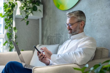 Foto de Hombre emprendedor sonriente con el ordenador portátil sentado en el sofá escribiendo un mensaje de texto en el teléfono inteligente - Imagen libre de derechos