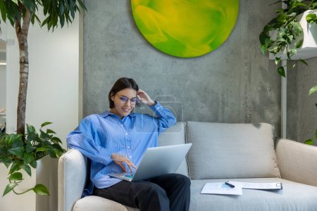Téléchargez les photos : Agréable jeune employée de bureau assise sur le canapé lisant quelque chose sur l'ordinateur portable - en image libre de droit