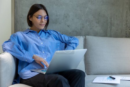 Téléchargez les photos : Calme concentré jeune employé à distance caucasien assis sur le canapé tapant sur l'ordinateur portable - en image libre de droit
