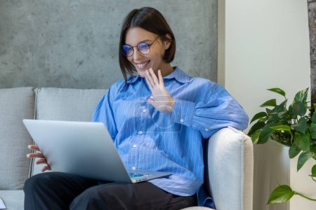 Téléchargez les photos : Jeune femme souriante assise sur le canapé saluant quelqu'un sur l'écran de l'ordinateur pendant l'appel vidéo - en image libre de droit