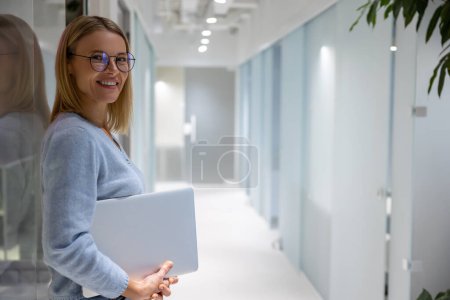 Photo for Contented young female corporate worker with the laptop in her hands leaning against the glass wall - Royalty Free Image