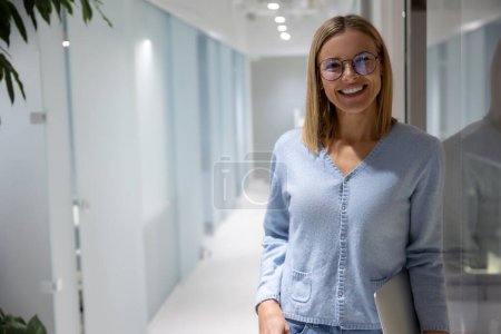 Photo for Merry female corporate worker with the laptop in the hand leaning against the glass wall - Royalty Free Image