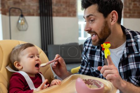 Foto de Padre barbudo alimentando al bebé a la hora de la comida, niño pequeño tratando de comer gachas o puré primera vez. - Imagen libre de derechos