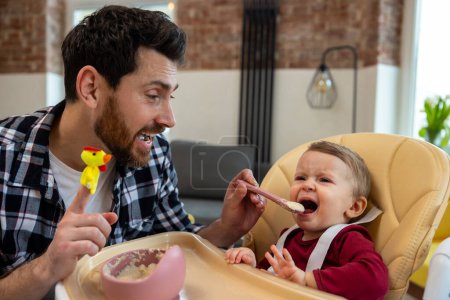 Photo for Little child care. Father feeding crying capricious infant daughter sitting at home in modern interior. - Royalty Free Image