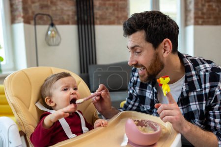 Téléchargez les photos : Petite garderie. Père nourrissant pleurer capricieuse petite fille assise à la maison dans l'intérieur moderne. - en image libre de droit