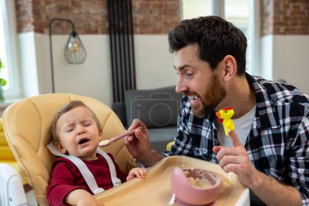 Photo for Little child care. Father feeding crying capricious infant daughter sitting at home in modern interior. - Royalty Free Image