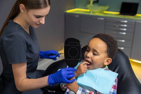 Téléchargez les photos : En dentisterie. Docteur donnant miroir au garçon à la peau foncée - en image libre de droit
