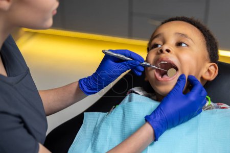 Téléchargez les photos : En dentisterie. Dentiste féminine travaillant avec un garçon à la peau foncée - en image libre de droit
