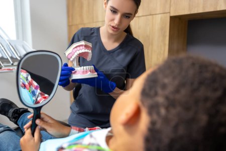 Foto de En odontología. Niño de piel oscura mirándose en el espejo mientras la doctora le muestra una maqueta de mandíbula - Imagen libre de derechos