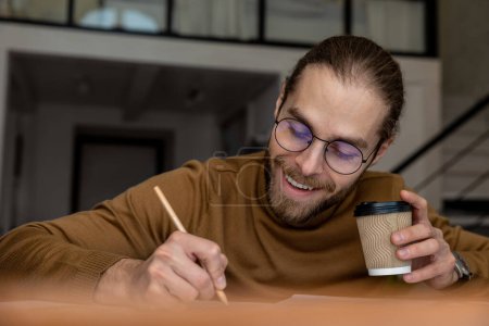 Junge professionelle männliche Büroangestellte schreiben Dokumente ein, trinken Kaffee zum Mitnehmen und sitzen an ihrem Arbeitsplatz zu Hause oder im Büro.