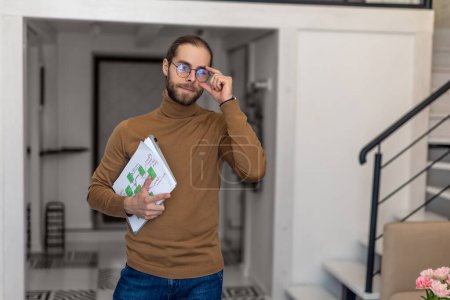 Téléchargez les photos : Bel homme dans des lunettes avec barbe debout tenant graphique en papier à l'intérieur de la maison. - en image libre de droit