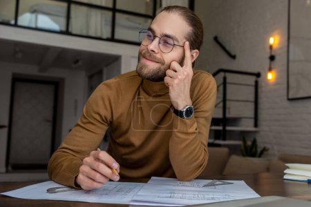 Photo for Portrait of pensive bearded young engineer architect in glasses sitting at the table and creating blueprint thinking about new project. - Royalty Free Image