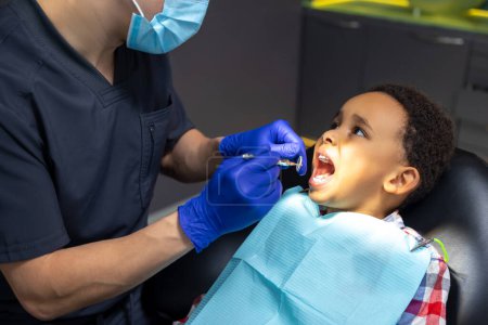 Foto de En odontología. Niño de piel oscura mirando asustado mientras el médico revisa sus dientes - Imagen libre de derechos