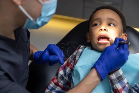 Téléchargez les photos : À la dentisterie. Garçon à la peau foncée regardant effrayé tandis que le médecin vérifie ses dents - en image libre de droit