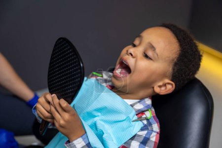 Foto de En los dentistas. Niño de piel oscura mirando sus dientes en el espejo - Imagen libre de derechos