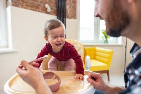 Téléchargez les photos : Tout-petit mignon bébé fille pleurer et ne veulent pas manger de la nourriture de son père qui se nourrit avec une cuillère. - en image libre de droit