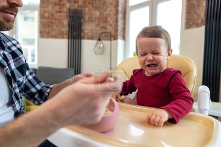 Téléchargez les photos : Tout-petit mignon bébé fille pleurer et ne veulent pas manger de la nourriture de son père qui se nourrit avec une cuillère. - en image libre de droit