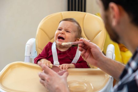 Téléchargez les photos : Père méconnaissable nourrir pleurer tout-petit fille tenant cuillère enfant manger purée. - en image libre de droit