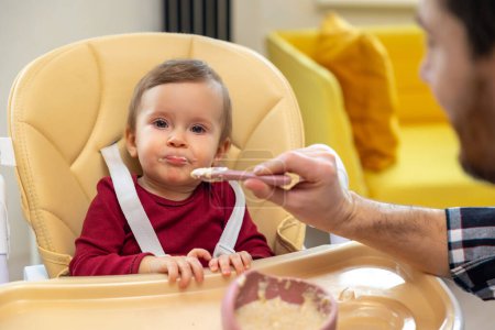 Foto de Padre irreconocible alimentando a la pequeña hija sentada en silla alta con puré o gachas de avena por cuchara en casa. - Imagen libre de derechos