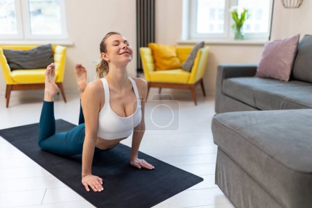 Téléchargez les photos : Jeune femme en forme en vêtements de sport pratiquant le yoga sur tapis d'exercice à l'intérieur de la maison. - en image libre de droit
