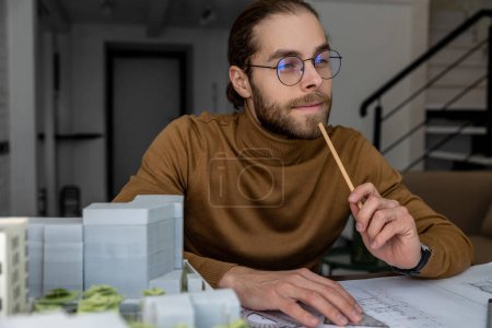 Téléchargez les photos : Portrait de coûteux jeune ingénieur barbu dans des lunettes assis à la table et de créer un plan de réflexion sur un nouveau projet. - en image libre de droit