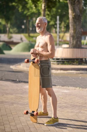 Téléchargez les photos : Homme mûr souriant tenant longboard dans un parc de la ville, mode de vie sain actif. - en image libre de droit