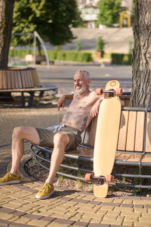 Téléchargez les photos : Satisfait homme mature avec longboard assis sur le banc dans un parc de la ville - en image libre de droit