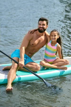 Téléchargez les photos : Souriant bel homme avec sa fille assis sur la planche de surf dans l'eau profiter des vacances passer du temps ensemble à la station. - en image libre de droit