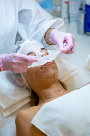 Téléchargez les photos : Cosmétologue méconnaissable enlever masque de feuille hydratante sur le visage féminin, soins de la peau et traitement de beauté. - en image libre de droit