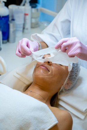 Téléchargez les photos : Cosmétologue méconnaissable enlever masque de feuille hydratante sur le visage féminin, soins de la peau et traitement de beauté. - en image libre de droit