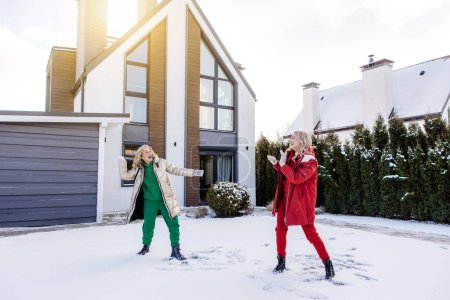 Photo for Smiling cheerful stylish beautiful blonde mature Caucasian woman throwing a snowball at her joyous daughter - Royalty Free Image