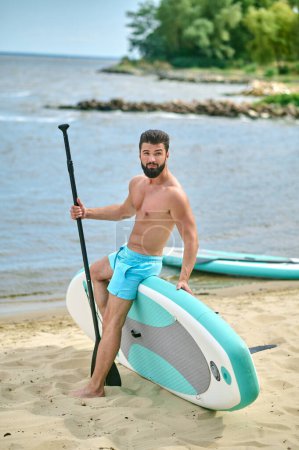 Téléchargez les photos : Beau sportif debout avec planche à pagaie sur une plage de sable près de la mer, profitant de vacances, mode de vie actif. - en image libre de droit