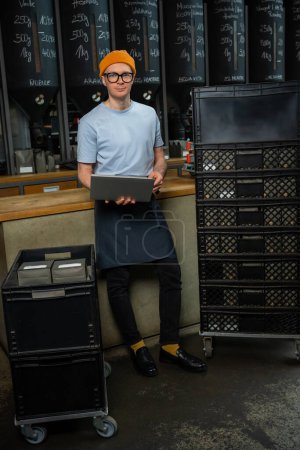 Foto de Barista guapo que trabaja en la cafetería, acepta nuevo producto, comprueba la cantidad de café, utilizando el portátil - Imagen libre de derechos