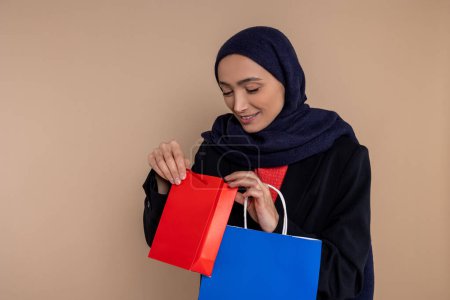 Photo for Joyful Muslim woman in hijab looking inside shopping bags isolated over beige background. - Royalty Free Image