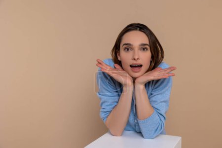 Photo for Surprised woman in casual clothing with hands under chin saying wow being astonished isolated over beige background. - Royalty Free Image