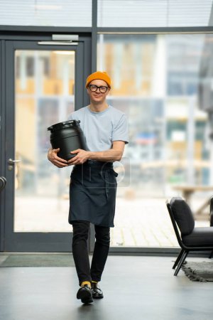 Téléchargez les photos : Jeune barista hipster portant chapeau, lunettes et tablier portant tonneau se préparant au travail dans un café. - en image libre de droit